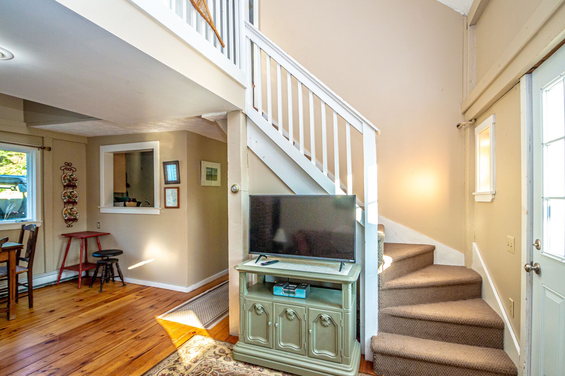 Interior with stairs, TV on a cabinet, and a door on the right. Warm tones with wooden floors.