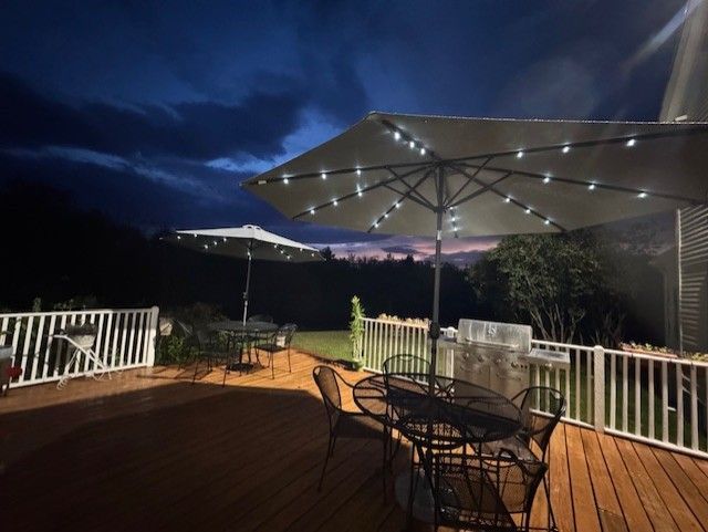 Wooden deck at dusk with lit umbrellas over outdoor tables, trees, and cloudy sky.