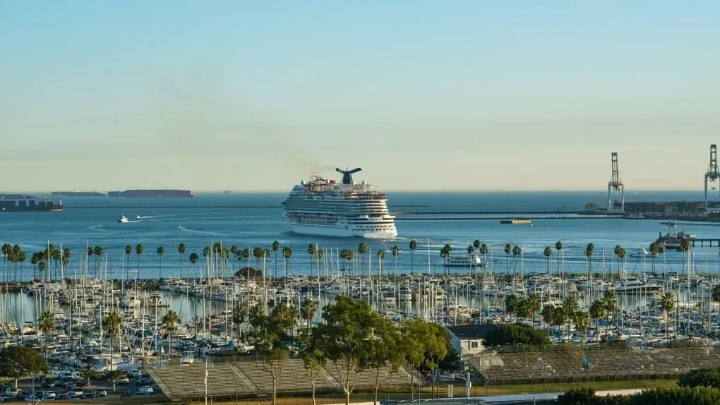 A large cruise ship sailing away from a harbor filled with smaller boats, palm trees, and industrial cranes at sunset.