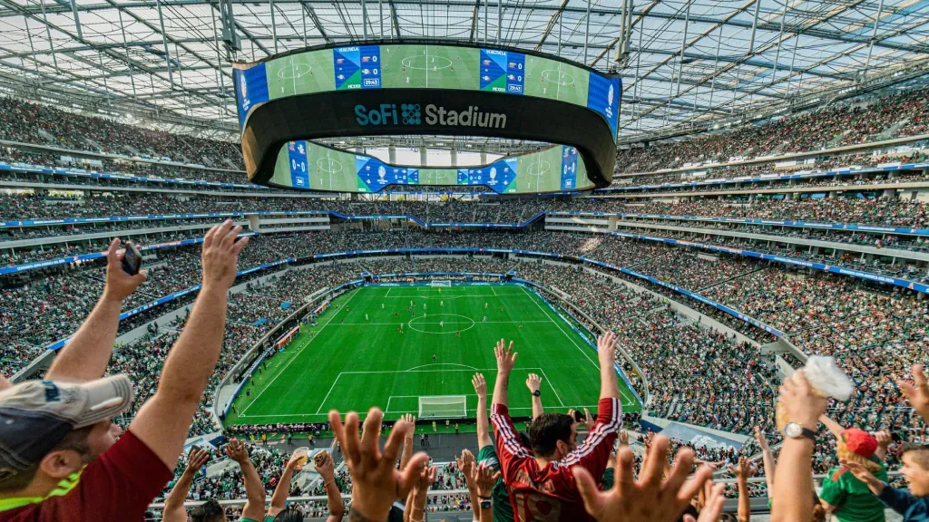 Fans with hands raised in celebration inside SoFi Stadium, overlooking a green soccer field under a large scoreboard.