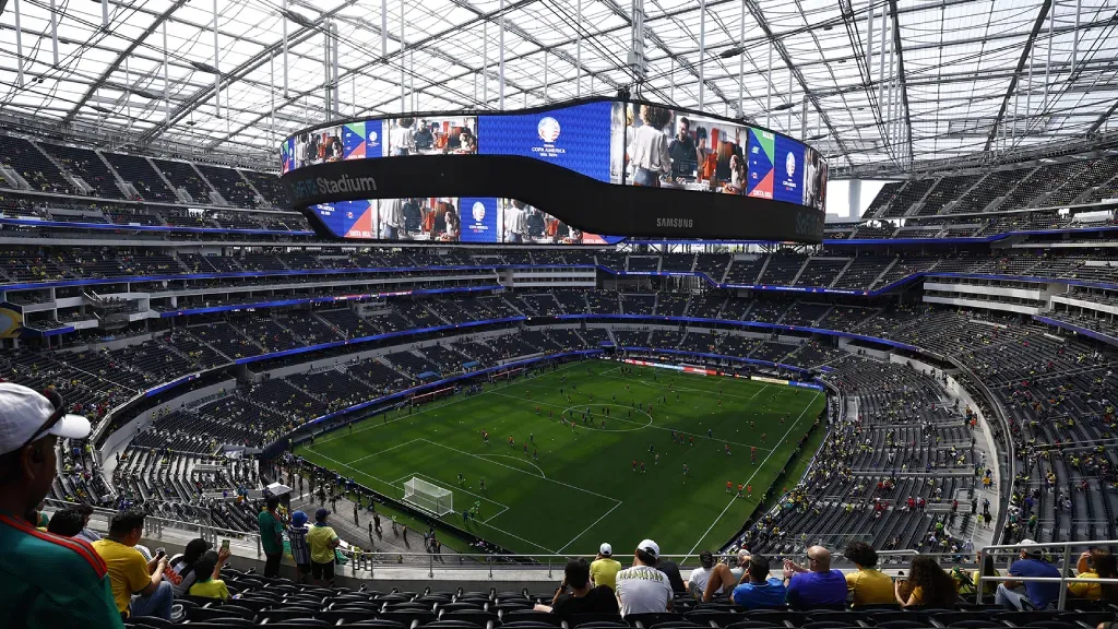 An elevated view of SoFi Stadium during a match, featuring a large oval scoreboard suspended above a green soccer field.