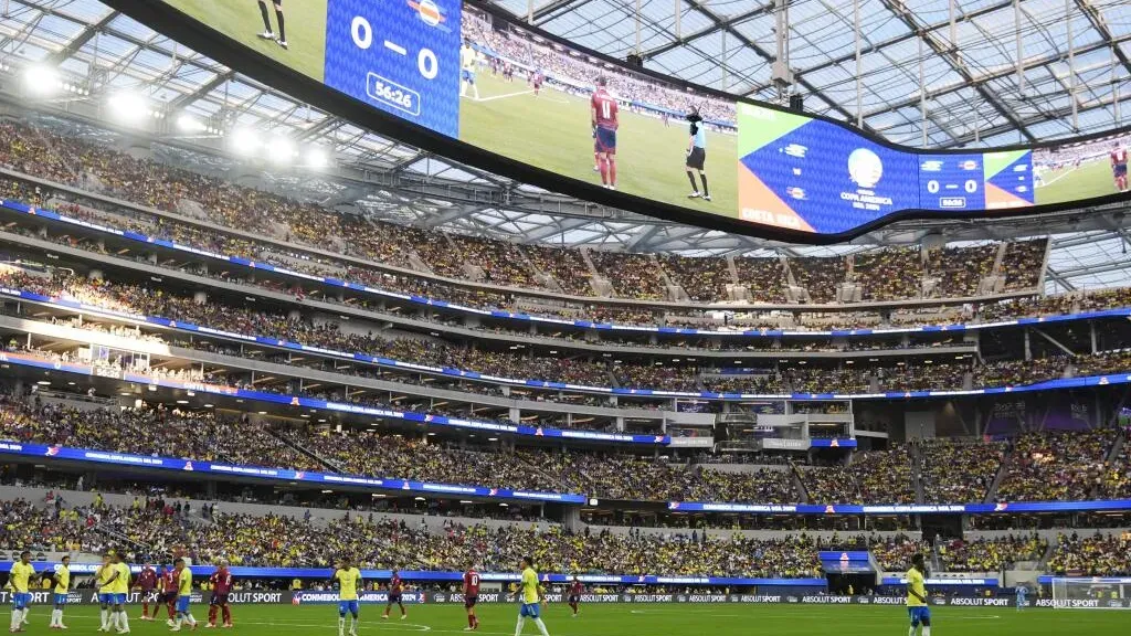 A large, packed stadium during a soccer match with a massive wrap-around screen displaying the 0-0 score above the field.