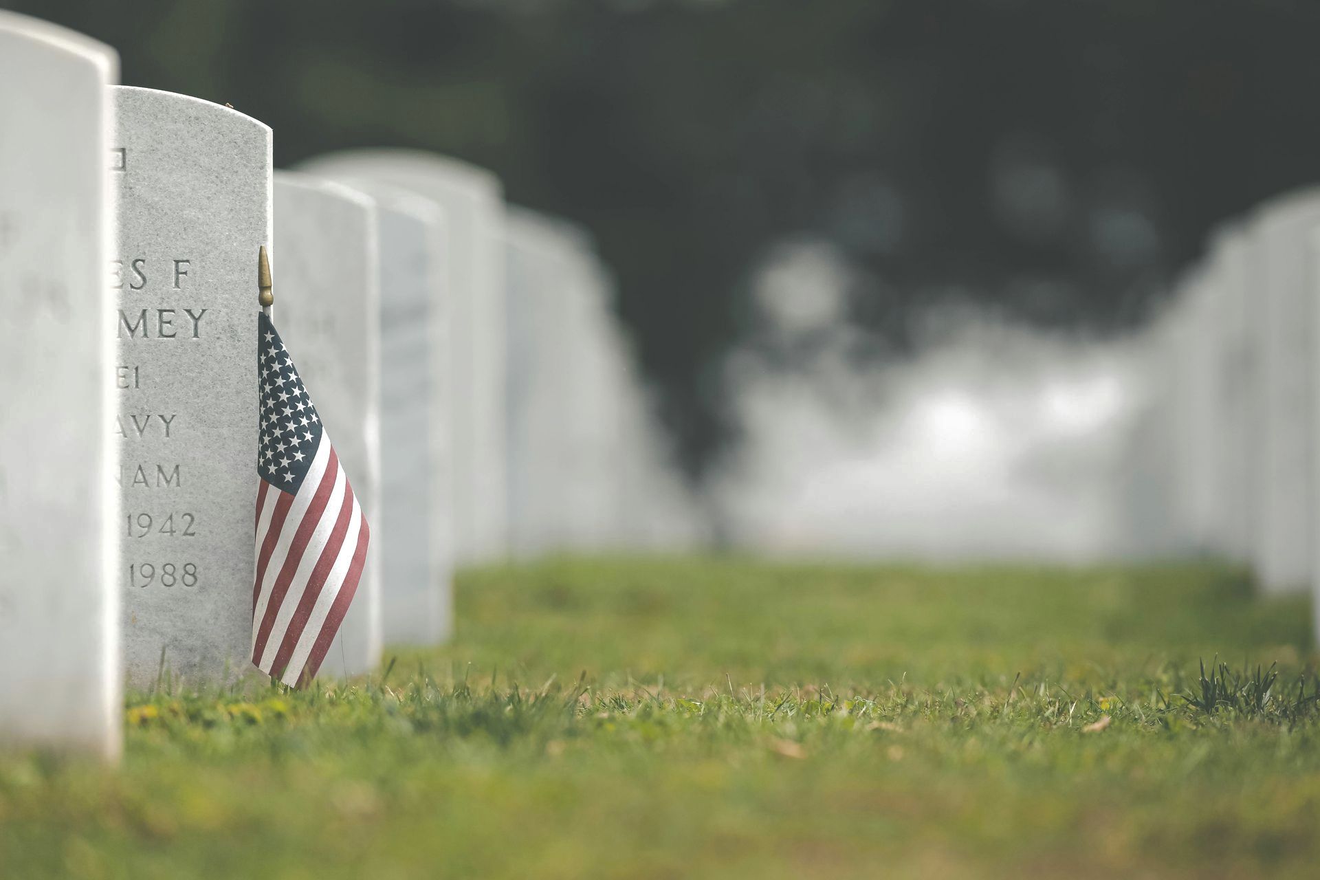 Row of white gravestones with a small American flag in the foreground on a misty cemetery lawn