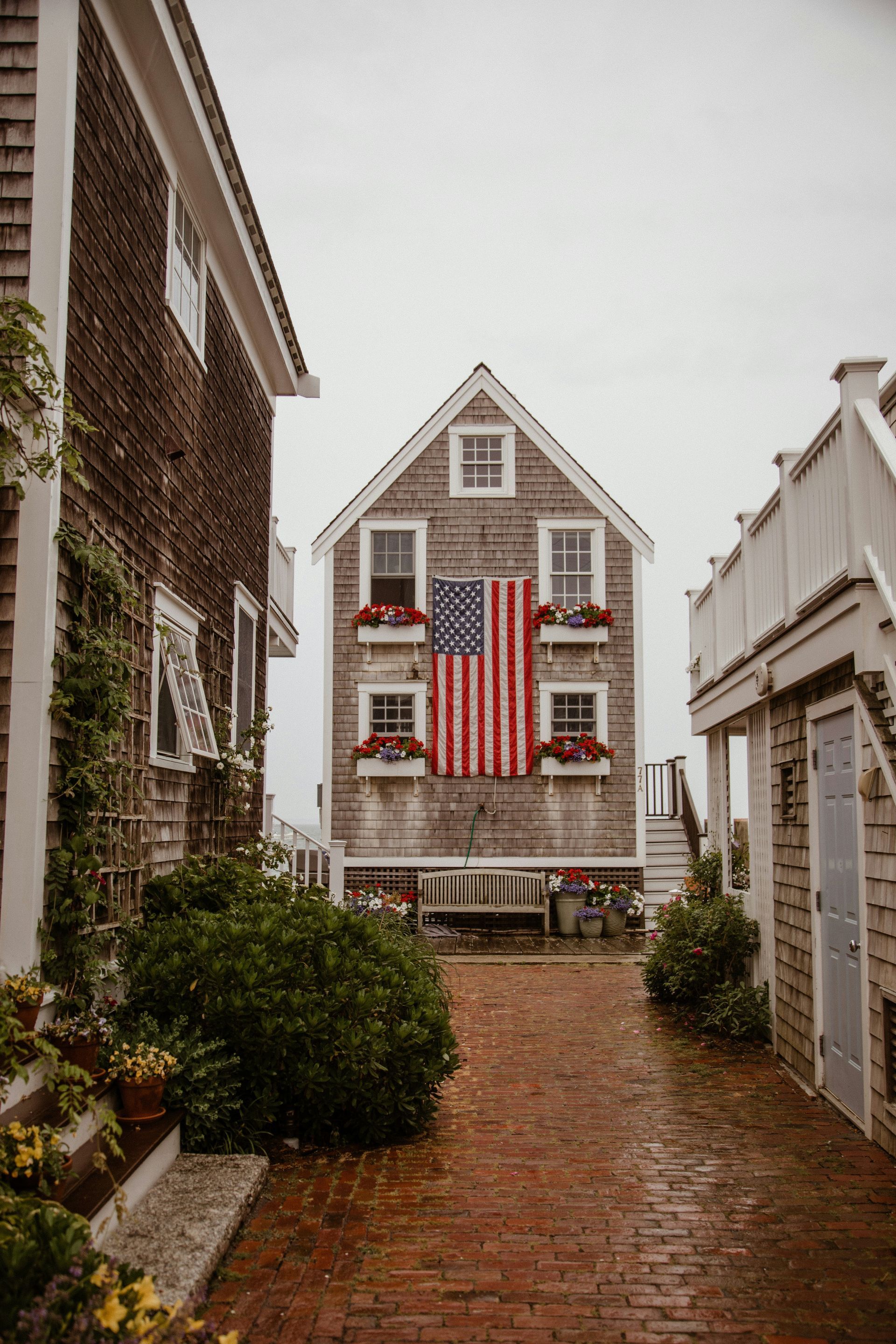 Narrow brick path leading to a gray house with red shutters and a large American flag flanked by white buildings