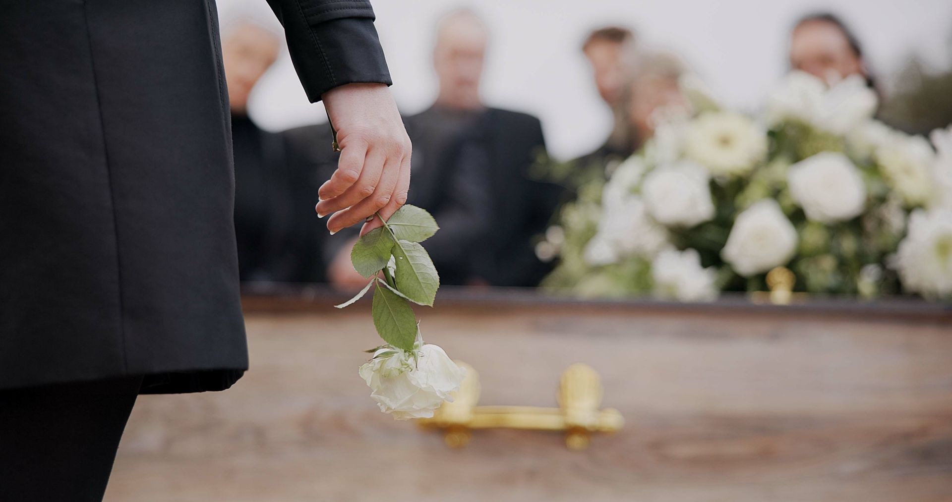 Hand placing white flowers on a coffin at a funeral, with mourners blurred in the background