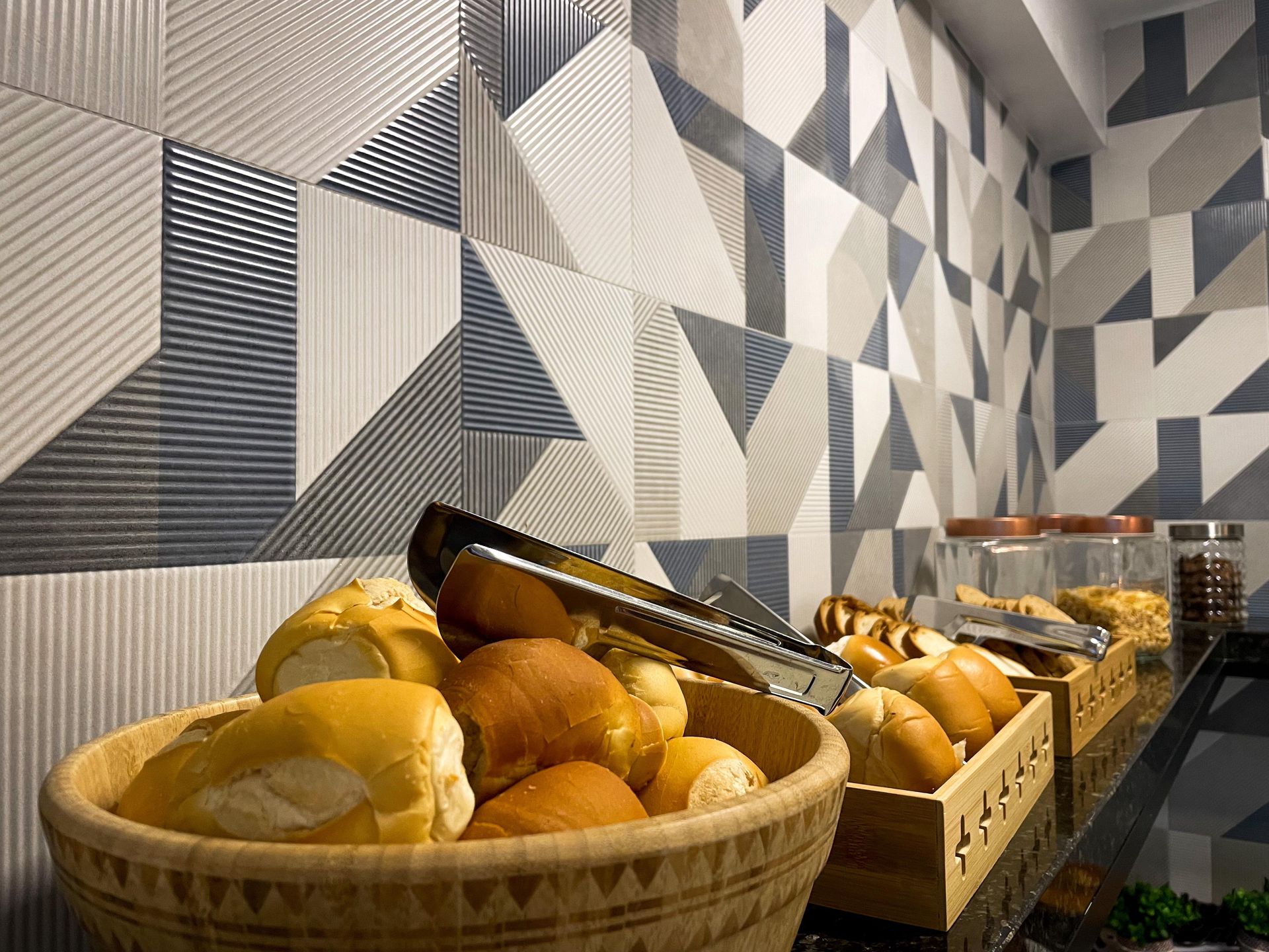 Duas cestas de madeira cheias de pãezinhos recém-assados estão sobre uma superfície encostada em uma parede de azulejos com padrões geométricos.