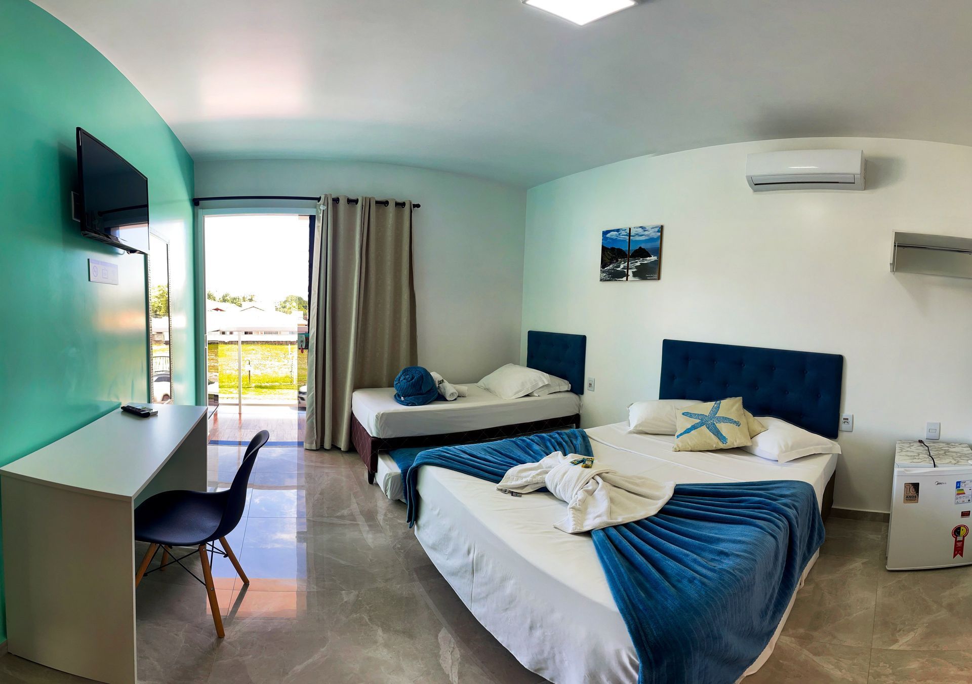 Um quarto de hotel com uma cama queen-size com um caminho de mesa azul e almofada em formato de estrela-do-mar, uma cama de solteiro, uma escrivaninha e uma varanda com vista.