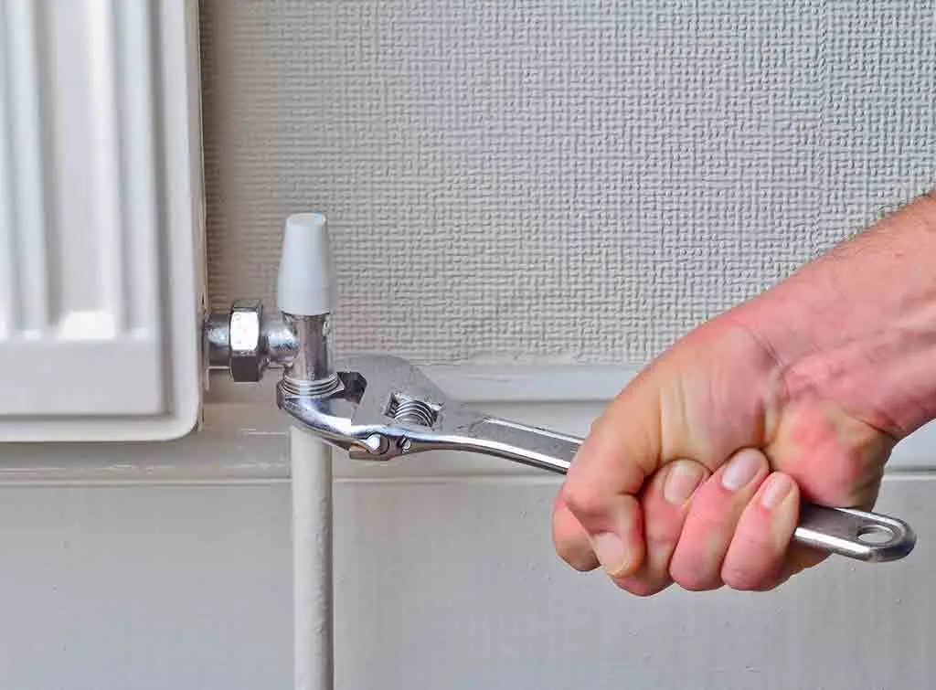 A hand uses an adjustable wrench to tighten a metal nut on the valve connected to a white radiator pipe.