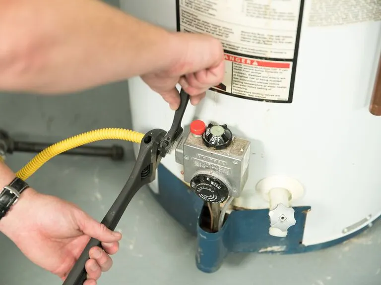 Hands using an adjustable wrench to tighten a yellow gas line connection on a water heater gas control valve.