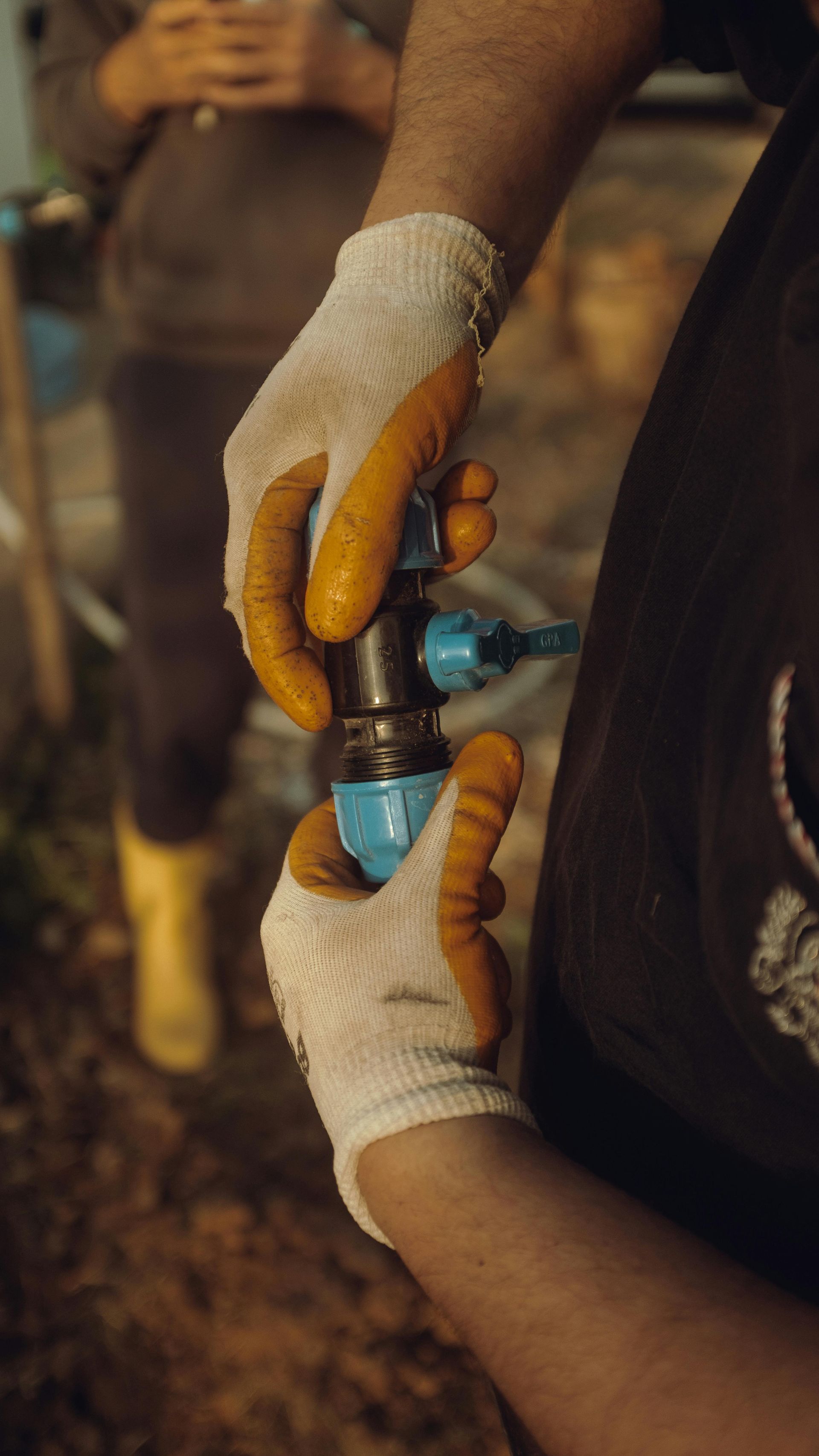 Hands in work gloves assemble a blue plastic plumbing fitting onto a metal pipe.
