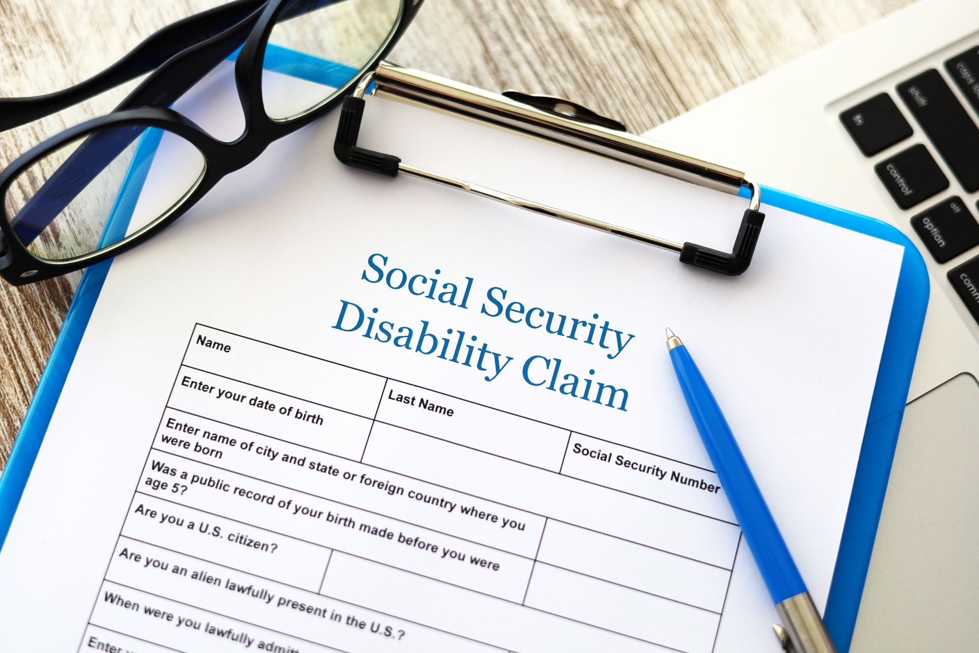 Disability claim form on clipboard with pen, glasses, and laptop.