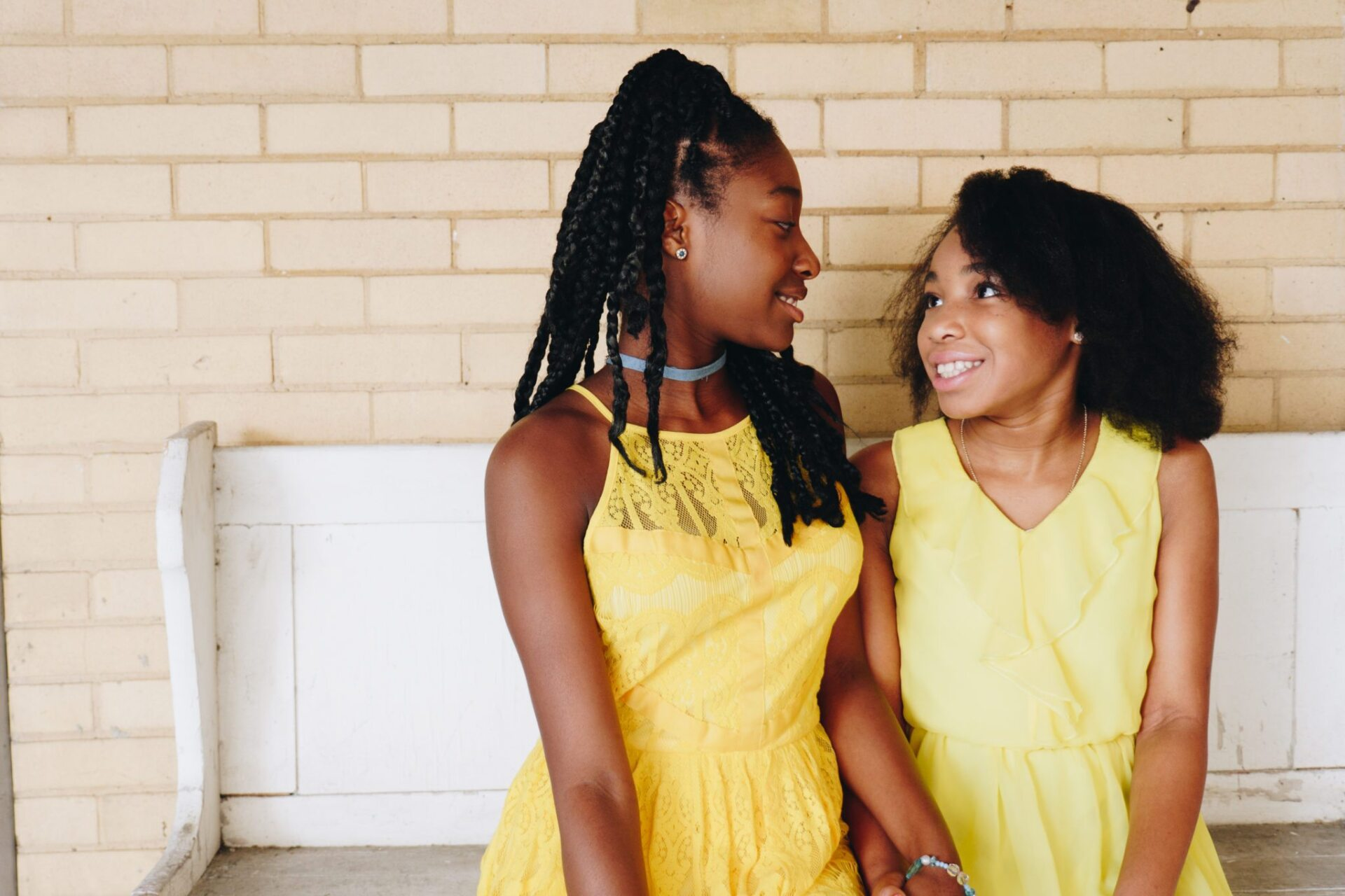 Two people in yellow dresses smiling at each other, sitting on a white bench, brick wall background.