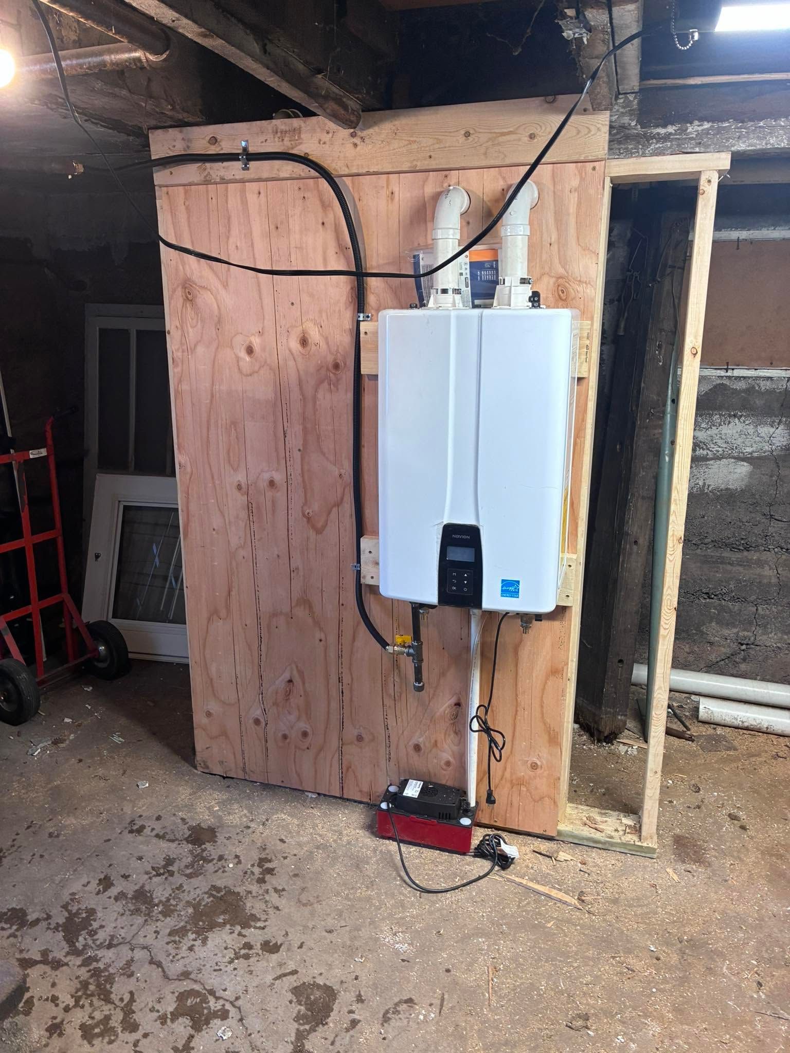 White wall-mounted boiler on plywood in a workshop, with exposed pipes and a red tool on the floor.