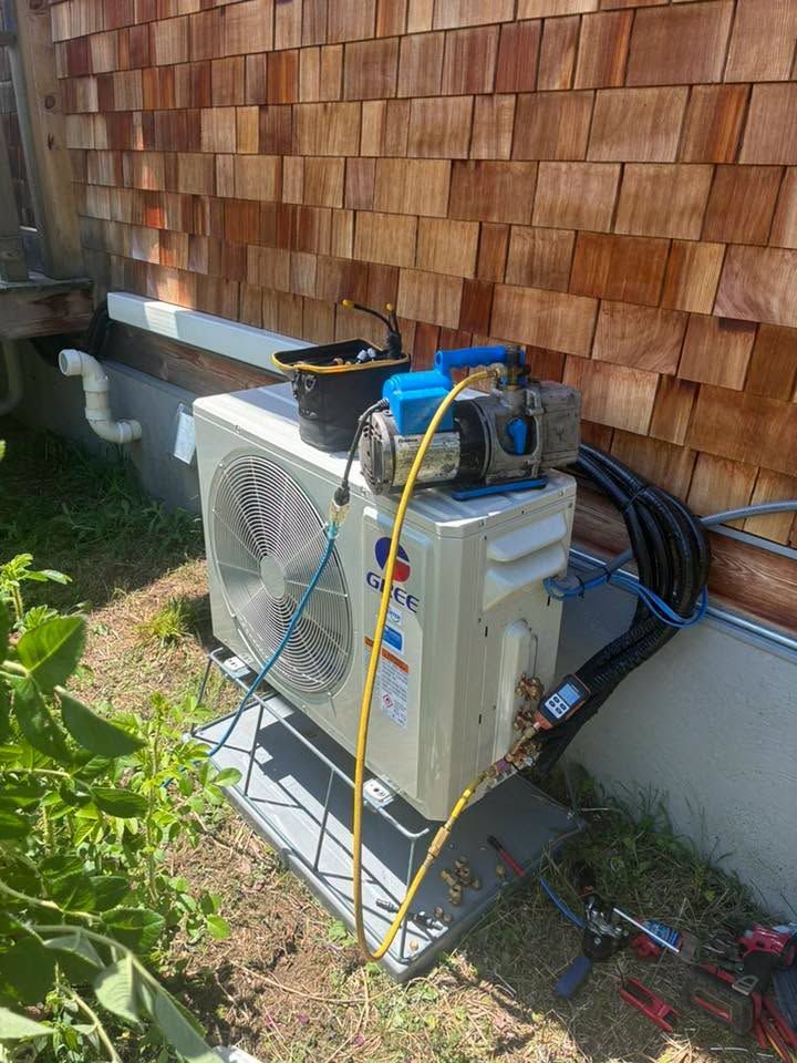 Outdoor HVAC unit beside a house wall, with tools and hoses on top and ground near the base.