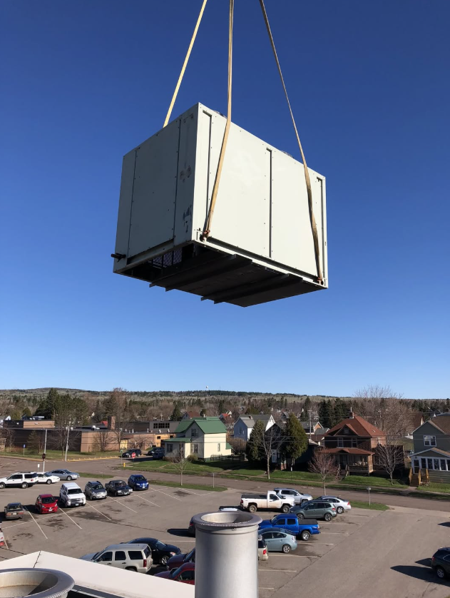 Large white box suspended by crane above a parking lot and neighborhood houses under a clear blue sky