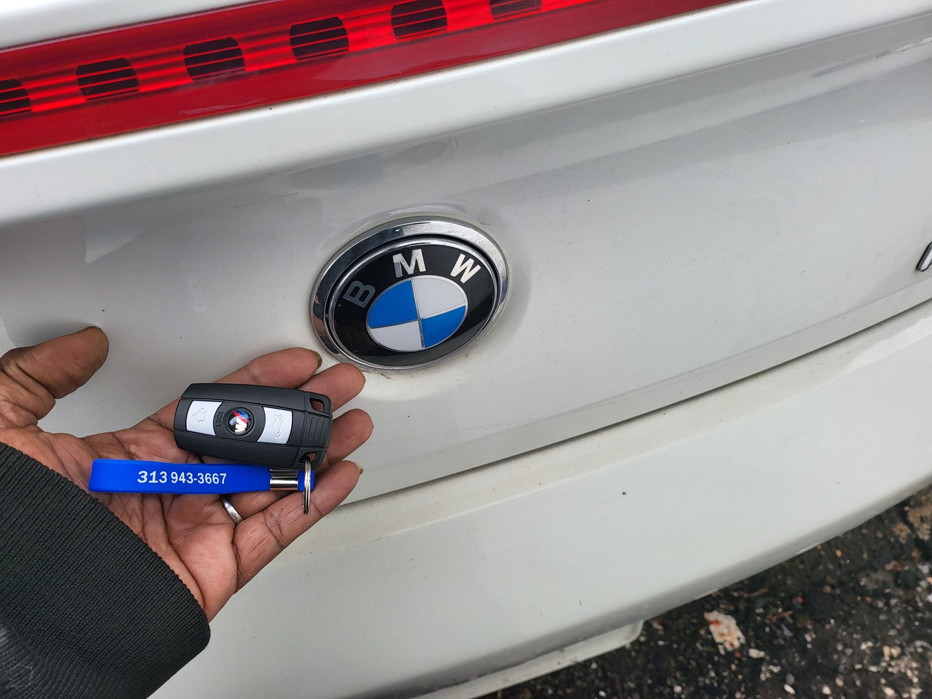 Hand holding car key and screwdriver near a white BMW emblem on a car's trunk.