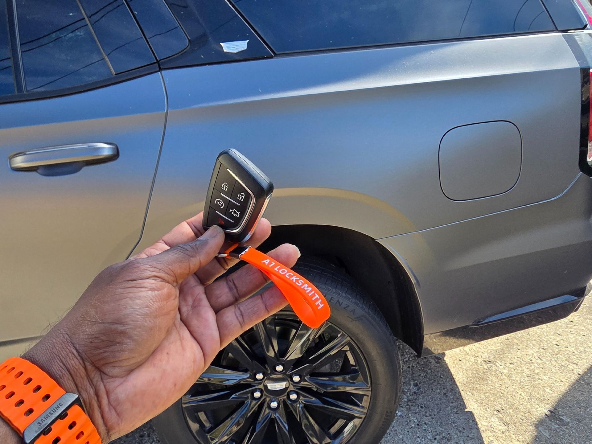 Hand holding car key fob near a gray SUV. Black wheels, orange wristband.