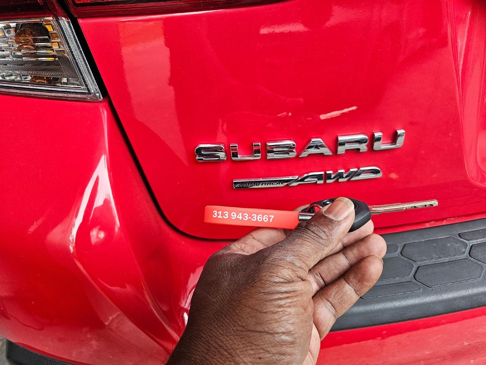 Hand holding a car key near the rear of a red Subaru. The car has a dent.