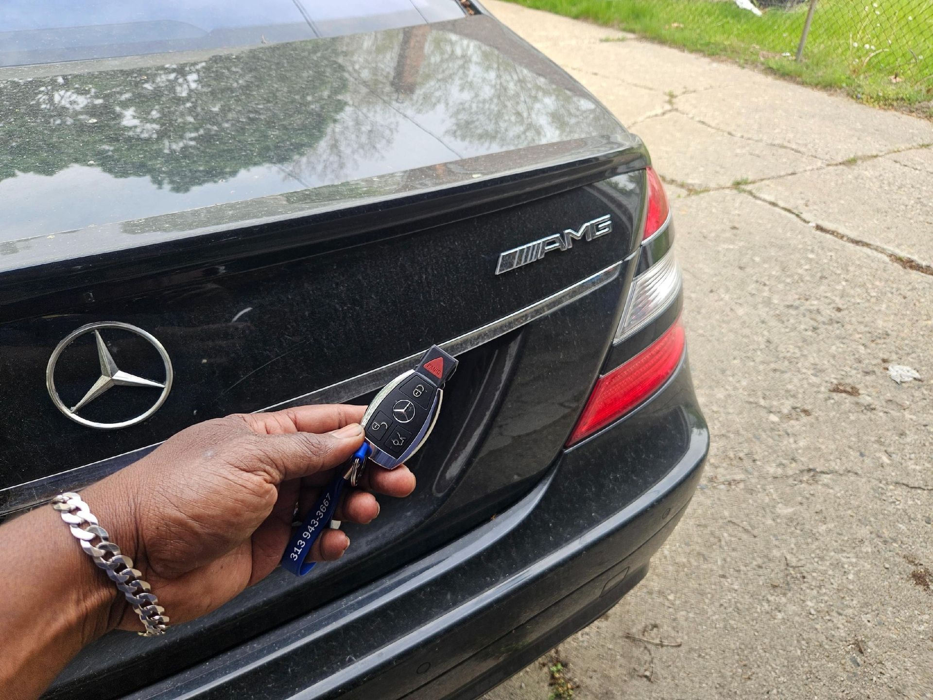 A hand with a silver bracelet holding a Mercedes-Benz key fob near the trunk of a black AMG car.