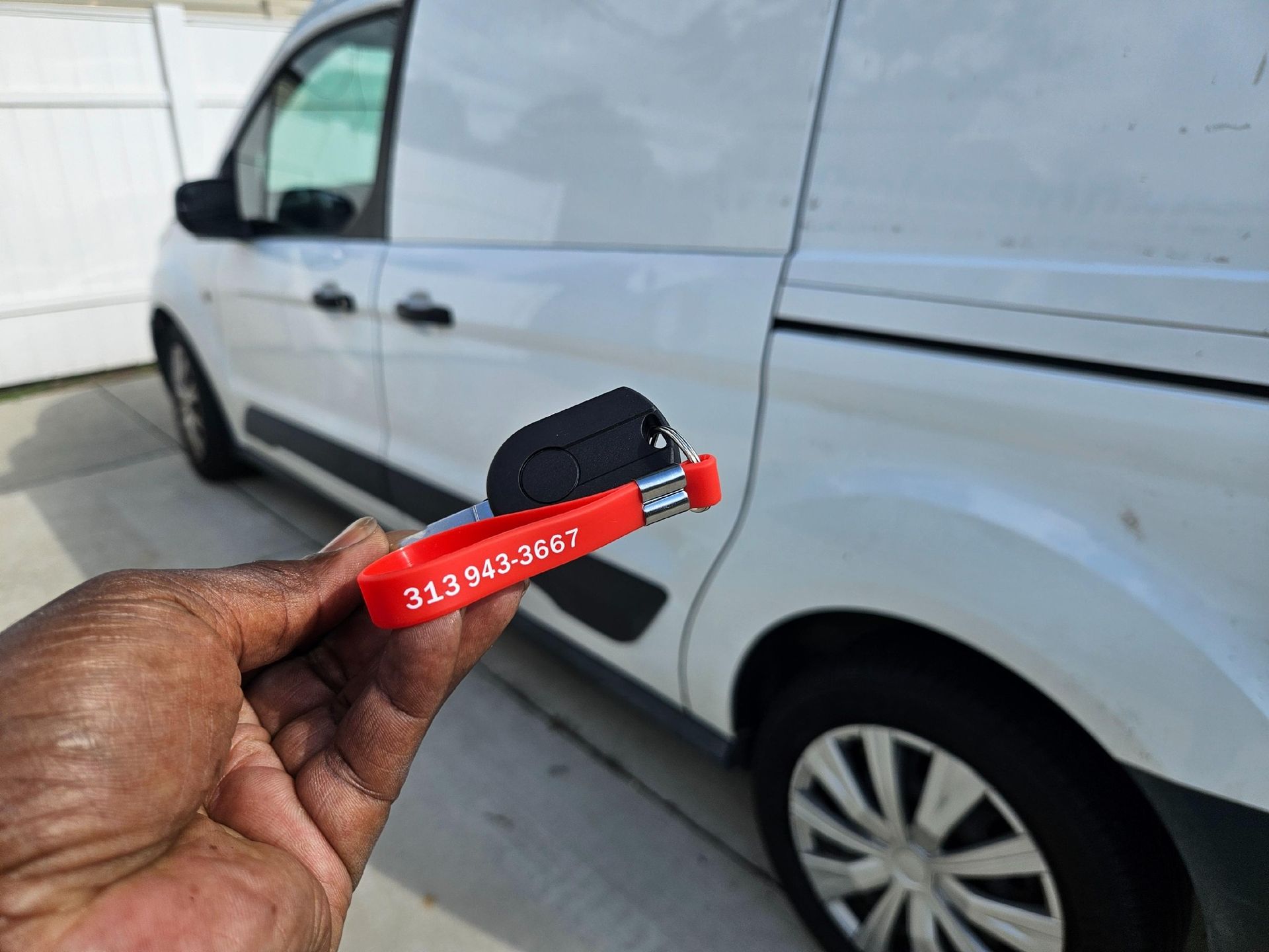 Hand holding a key fob and key with a red tag with a phone number, beside a white van.