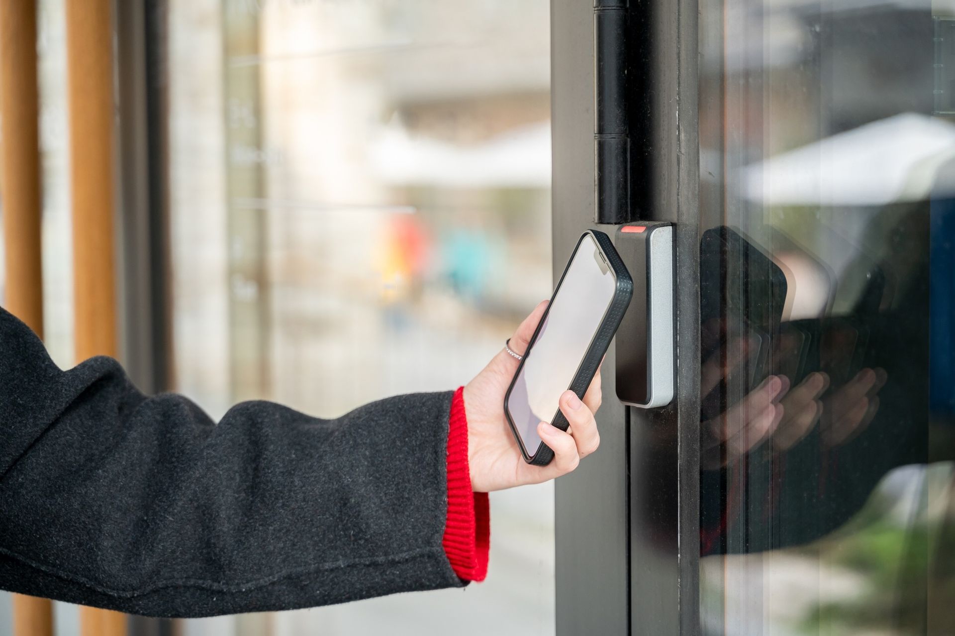 Person using a smartphone to unlock a door with a reader. Black coat, white phone, and black door frame.