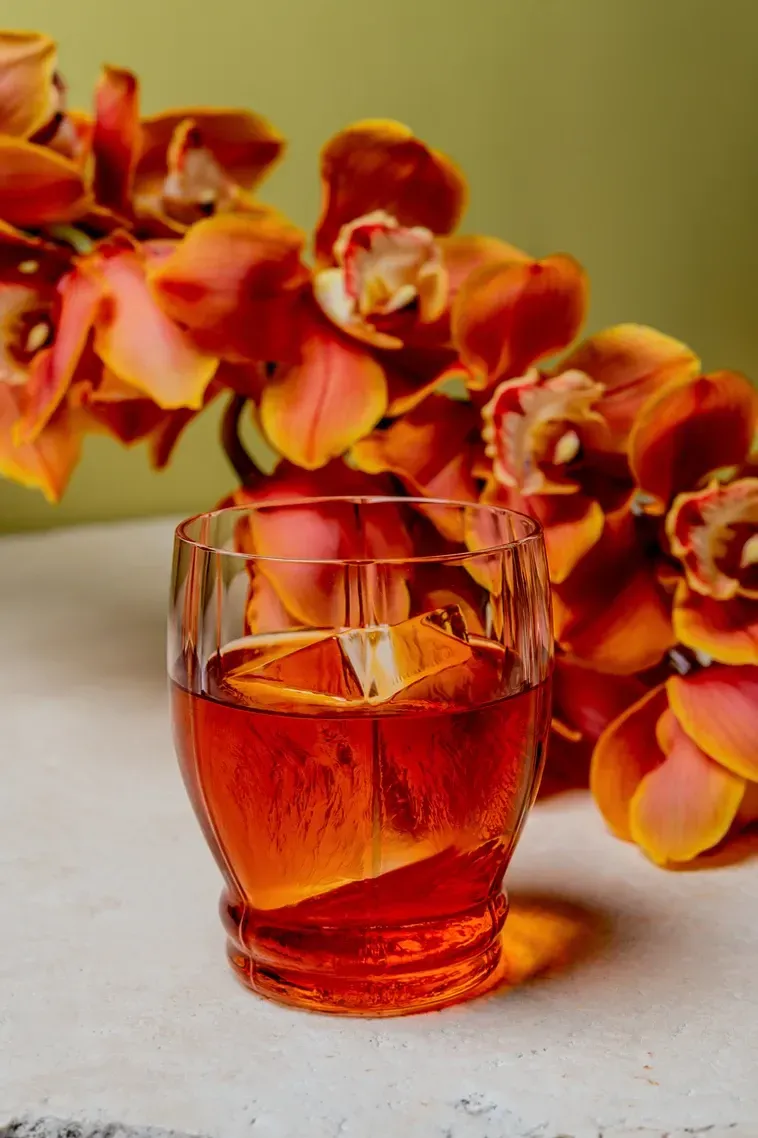 Cocktail with orange peel, in a glass with embossed pattern, next to orange orchids. Food Stylist Nadine Page.