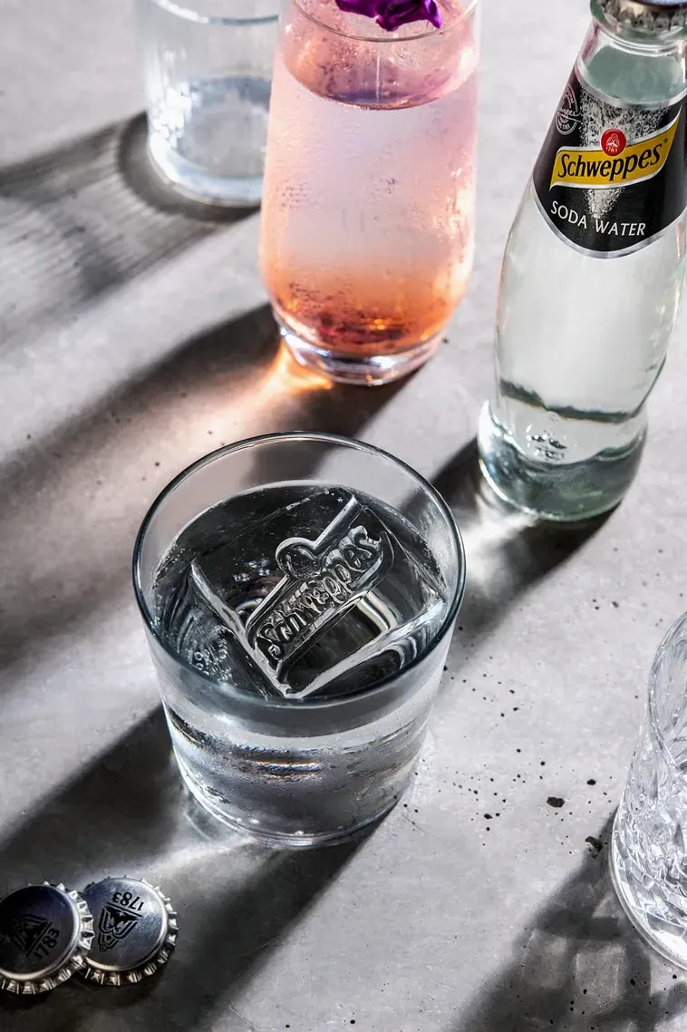 Cocktail with ice sculpture, pink drink, and Schweppes bottle on a table with bottle caps. Food Stylist Nadine Page.