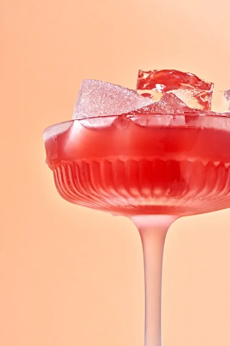 Pink cocktail in a frosted glass, garnished with a pink, sugar-coated cube against a peach background. Food Stylist Nadine Page.