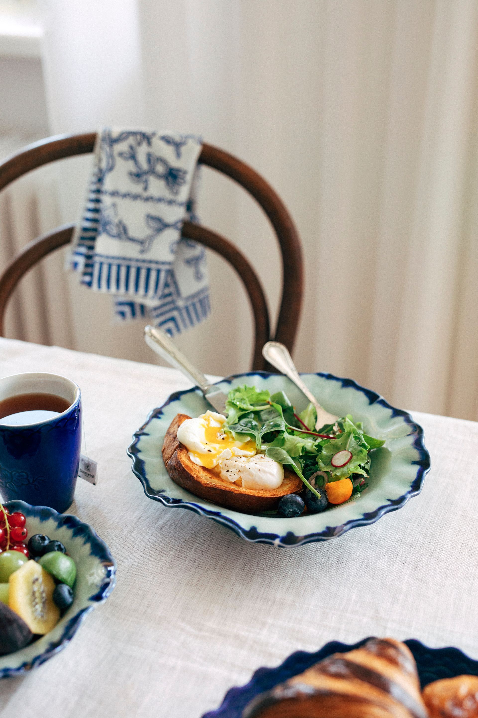 Breakfast table with plate of toast, egg, and greens; fruit in a blue bowl, cup of tea, towel on a chair. Food Stylist Nadine Page.