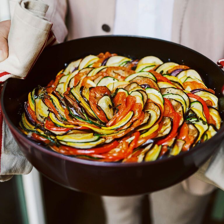 Ratatouille in a brown pan being held with oven mitts. Colorful vegetables arranged in concentric circles. Food Stylist Nadine Page.