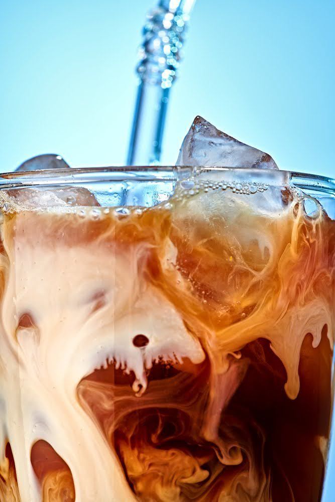 Iced coffee with milk swirling around ice cubes and a clear straw. Food Stylist Nadine Page.