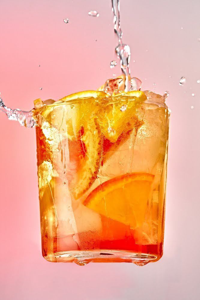 Orange cocktail with ice and citrus slices, being splashed with water against a gradient background. Food Stylist Nadine Page.