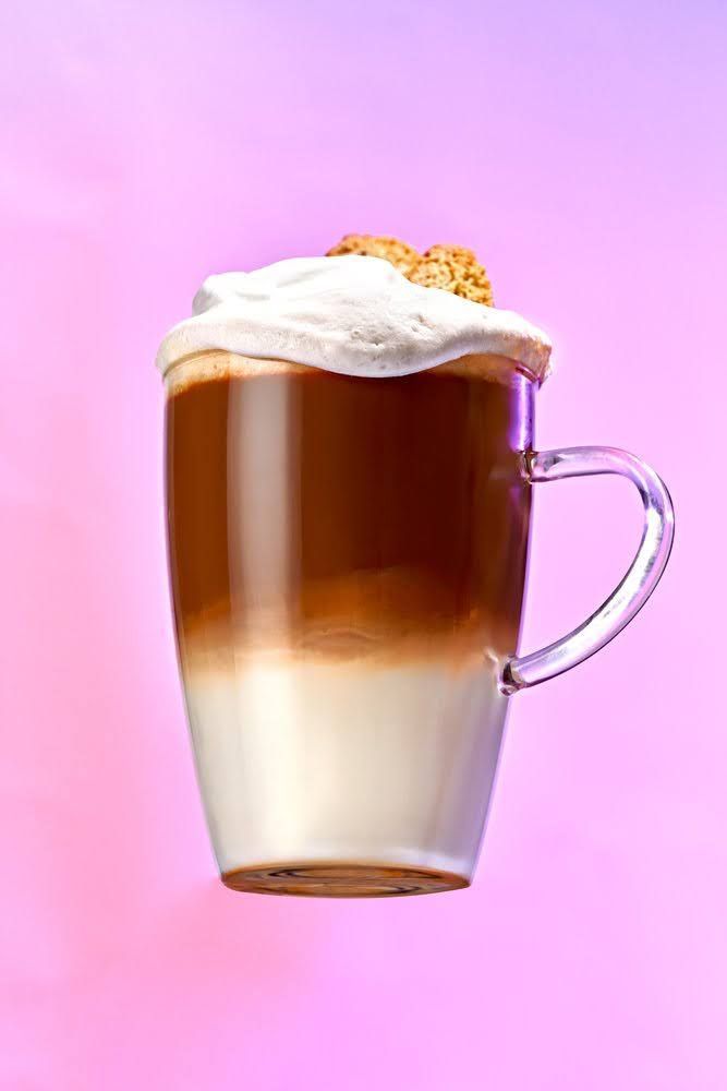 Coffee drink in layered colors, topped with whipped cream and a cookie, in front of a pink background. Food Stylist Nadine Page.