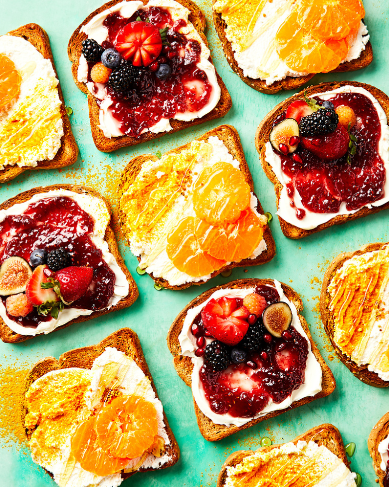 Toast slices topped with cream cheese and fruit, including berries and oranges, on a teal surface. Food Stylist Nadine Page.