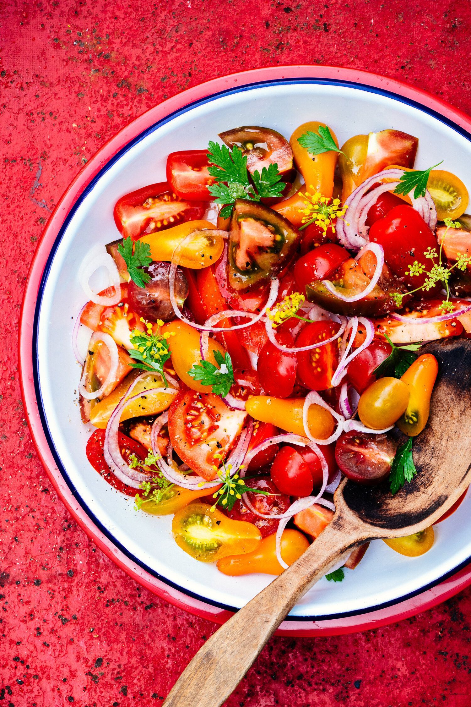 Colorful tomato salad in a white bowl with a wooden spoon on a red surface. Food Stylist Nadine Page.