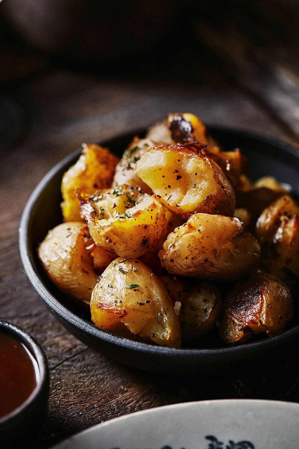 Roasted golden potatoes in a dark bowl, next to a small dish of sauce, on a wooden surface. Food Stylist Nadine Page.