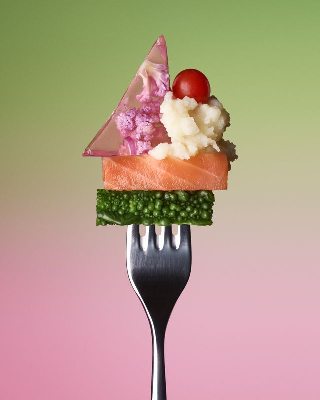Fork with stacked food: broccoli, salmon, cauliflower, tomato, and jelly, against pink and green background. Food Stylist Nadine Page.
