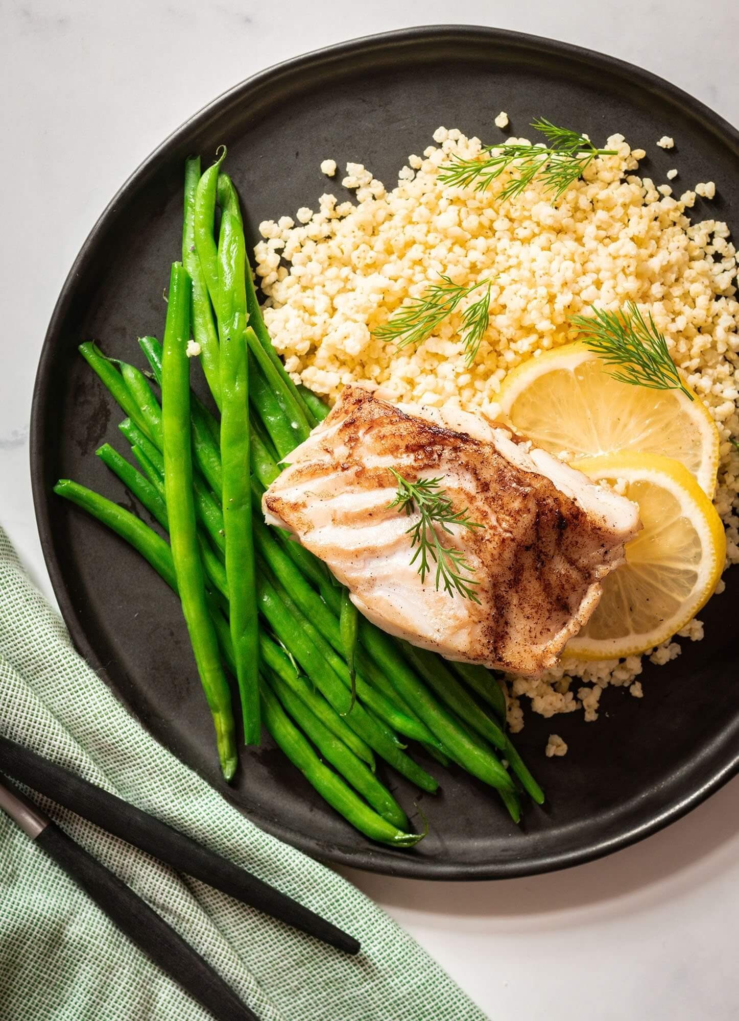 Black plate with seared fish, couscous, green beans, and lemon slices. Food Stylist Nadine Page.