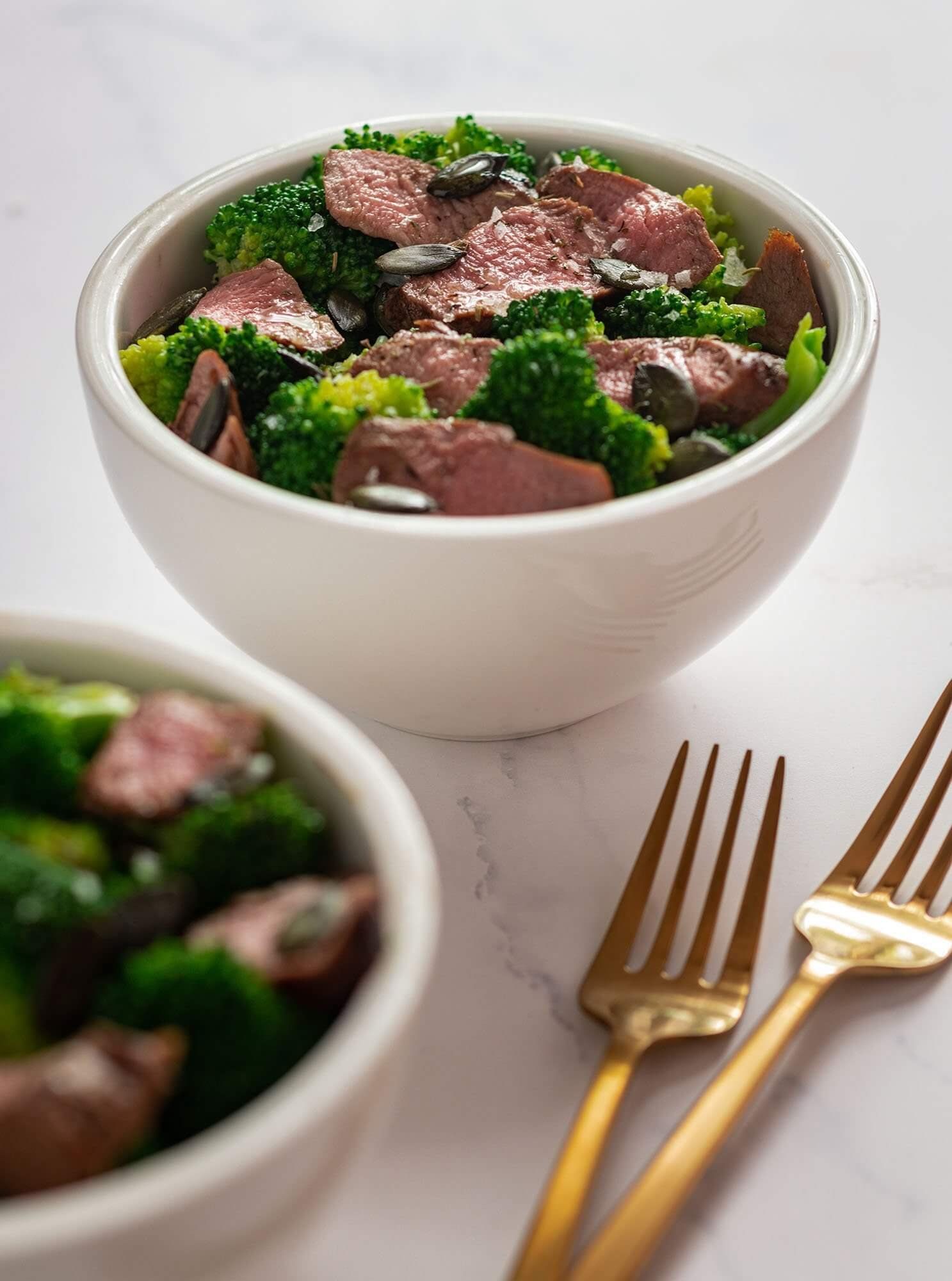 Bowls of steak and broccoli salad with gold forks on a white surface. Food Stylist Nadine Page.