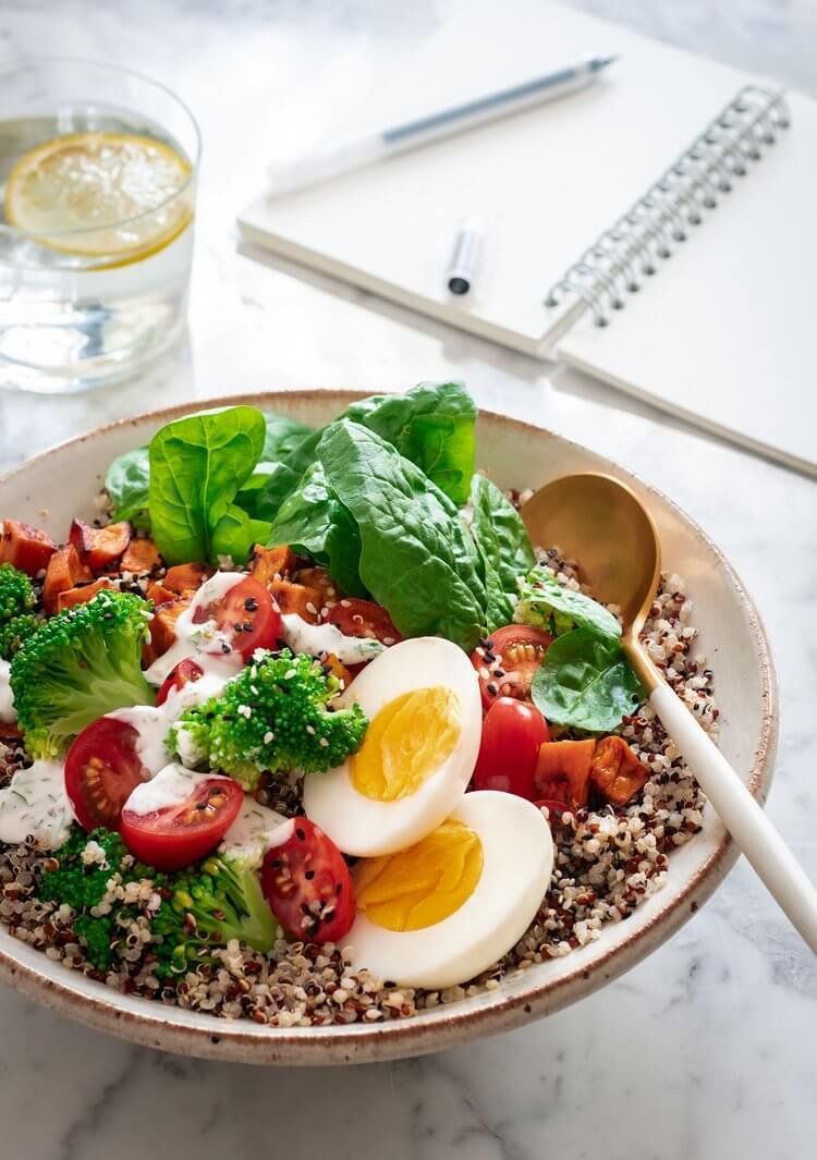 Healthy quinoa bowl with egg, broccoli, tomatoes, spinach, and dressing. Next to a glass of water, notebook, and pen. Food Stylist Nadine Page.