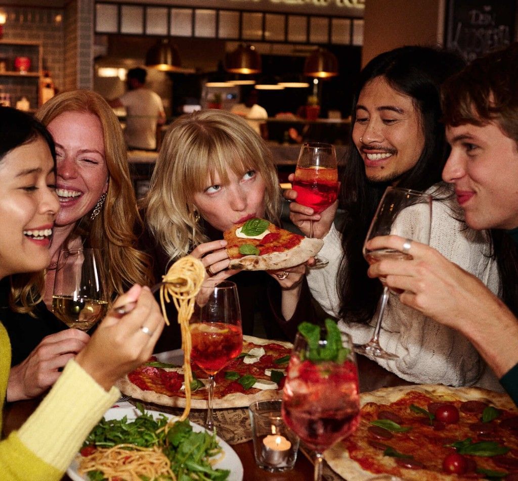 Friends at an Italian restaurant eating pizza and pasta, laughing, and enjoying drinks. Food Stylist Nadine Page.