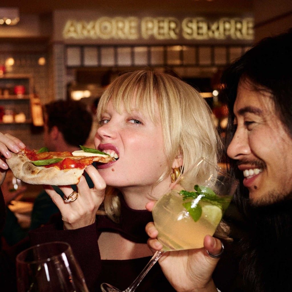 Woman eating pizza, man holding drink, at Italian restaurant with