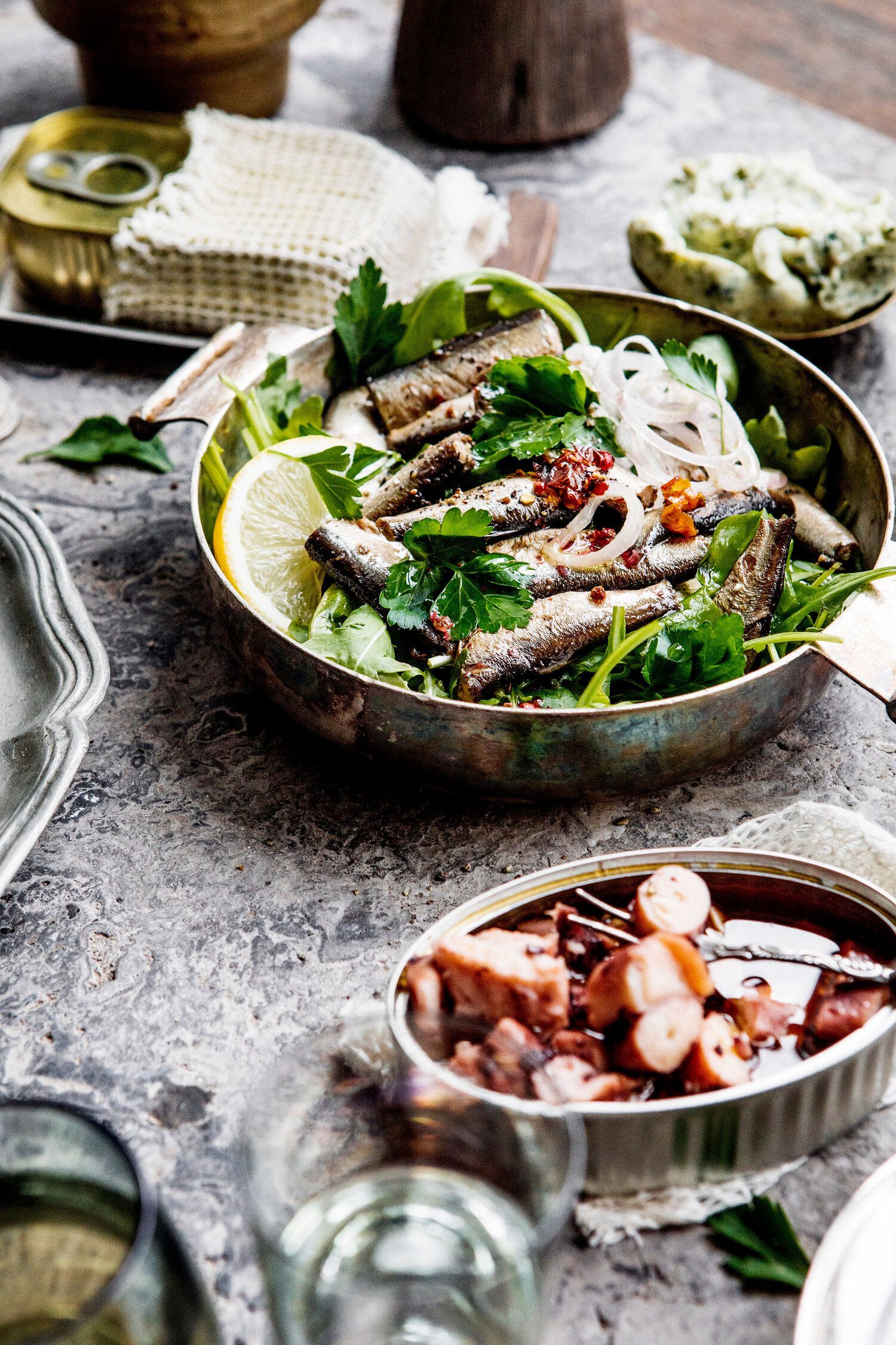 Close-up of canned sardines and octopus dishes with garnishes, on a rustic table. Food Stylist Nadine Page.