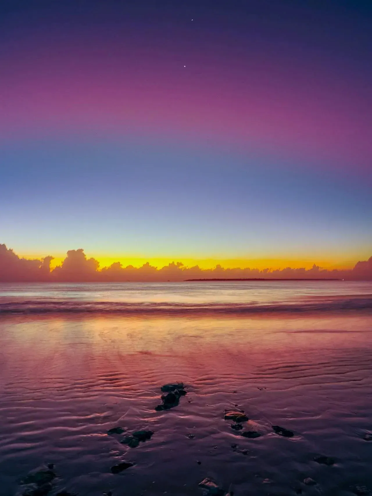 A sky of deep purple, pink, blue, and yellow/orange over a body of water, during sunrise.