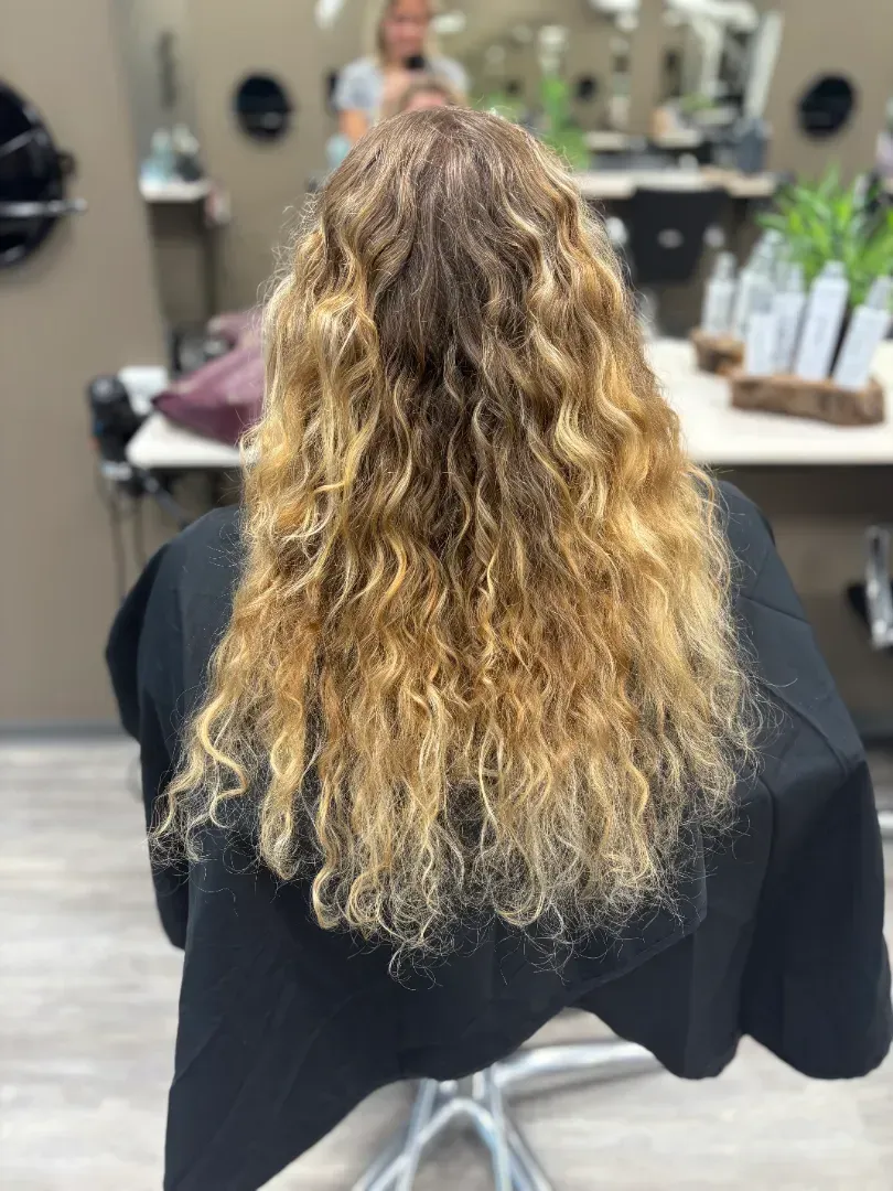 Woman with long, wavy, blonde hair in a salon chair, with a stylist in the background.