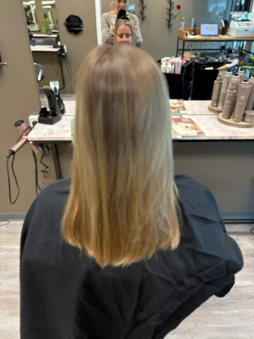Blonde-haired person in a salon, viewed from the back; hair cut and styled with a dark cape.