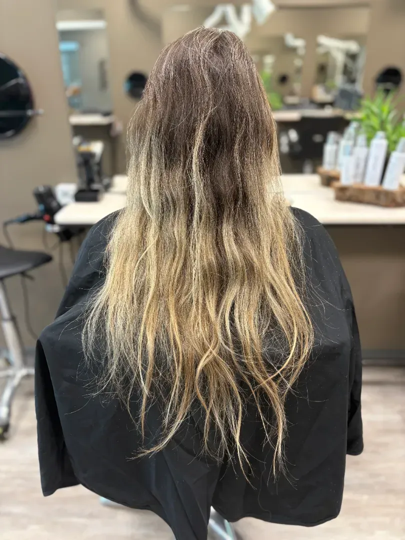 Woman with long, ombre hair in a salon chair, wearing a black cape. Hair is dark at the roots and blonde at the ends.