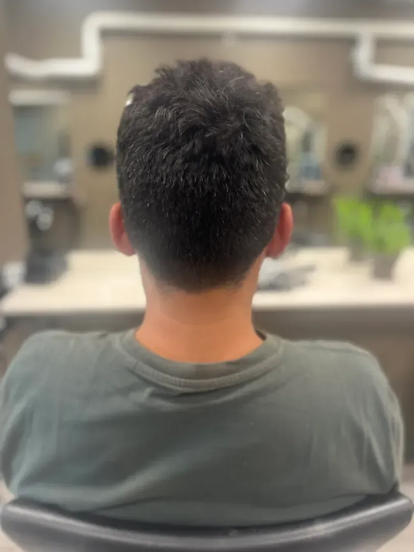 Person in a green shirt at a salon, facing away, with short, dark, textured hair and visible neckline.