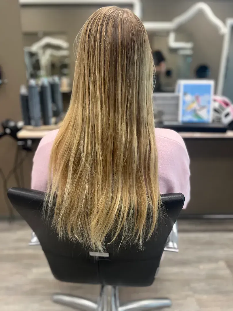 Woman with long, blonde hair sitting in a salon chair, seen from the back.