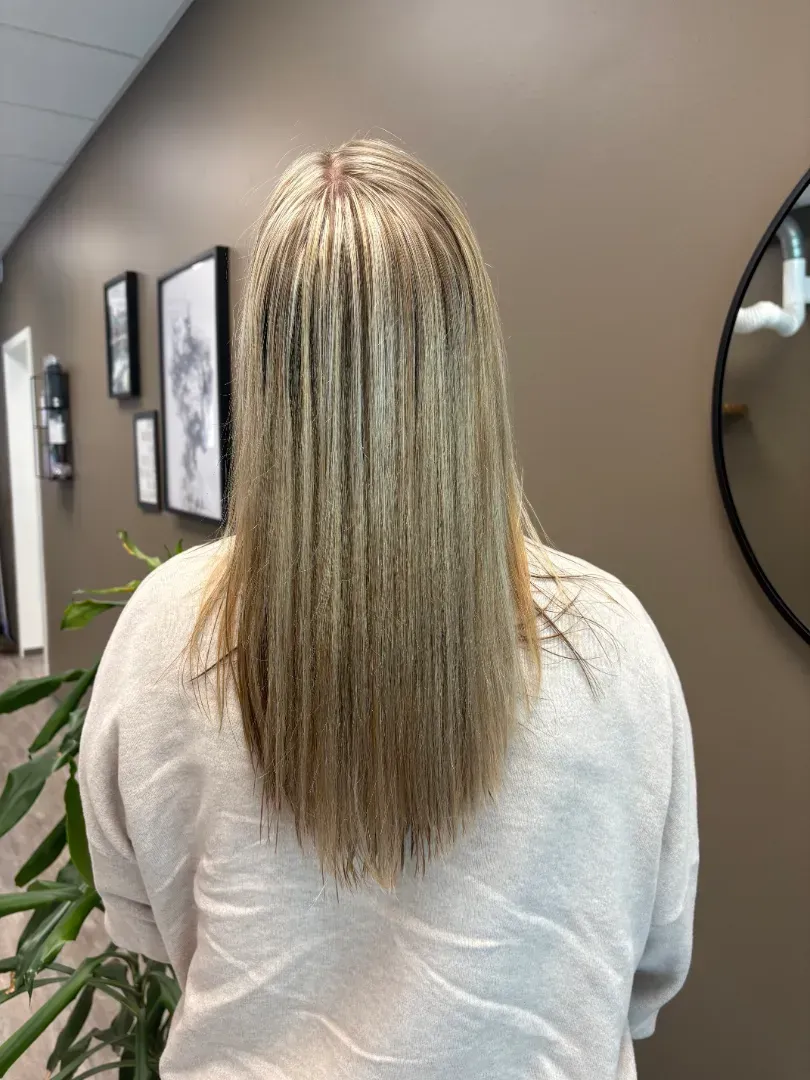 Woman with long blonde hair, facing away, in a salon. Neutral-toned wall, art, and mirror in the background.